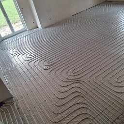 HYDRO Instal Mateusz Aseńczuk - Ułożone rury ogrzewania podłogowego na macie izolacyjnej w przestronnym, niewykończonym pomieszczeniu z dużym oknem, widok z góry.