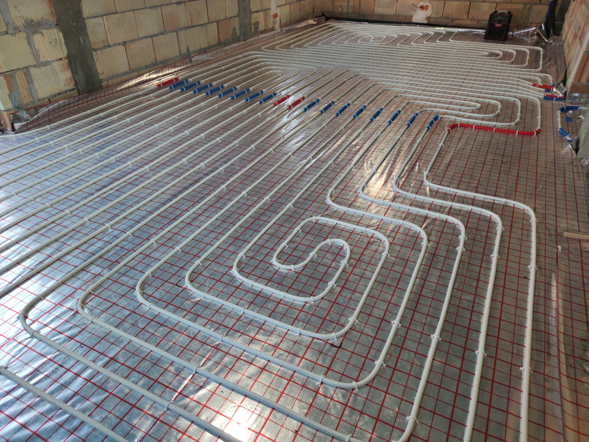 Białe rury systemu ogrzewania podłogowego ułożone spiralnie na folii z czerwoną siatką, widoczne elementy mocujące i fragment ściany z surowej cegły.