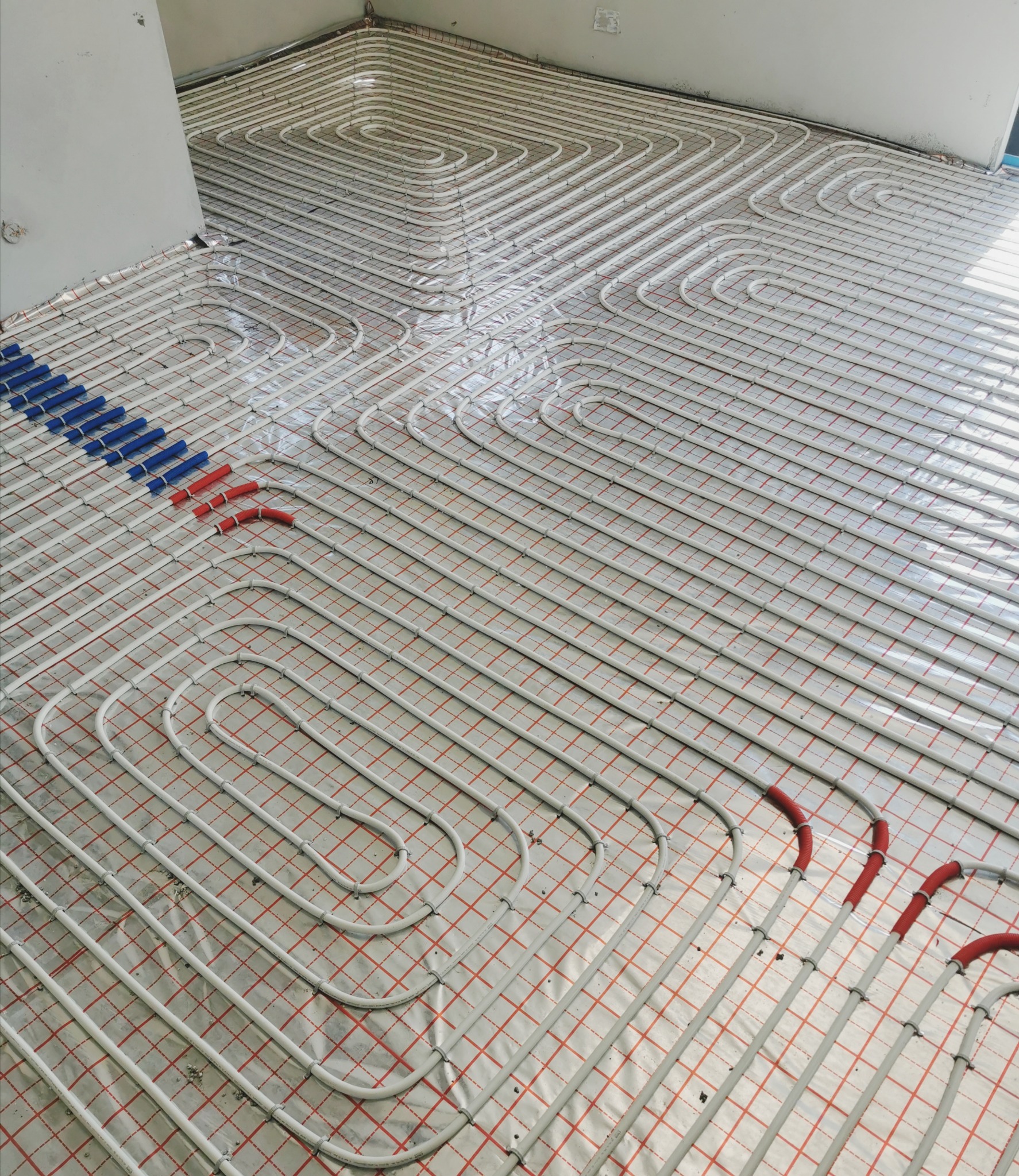 Białe rury systemu grzewczego ułożone w pętle na folii z czerwoną kratką, widoczne niebieskie i czerwone osłonki na rurach, przygotowanie do wylewki.