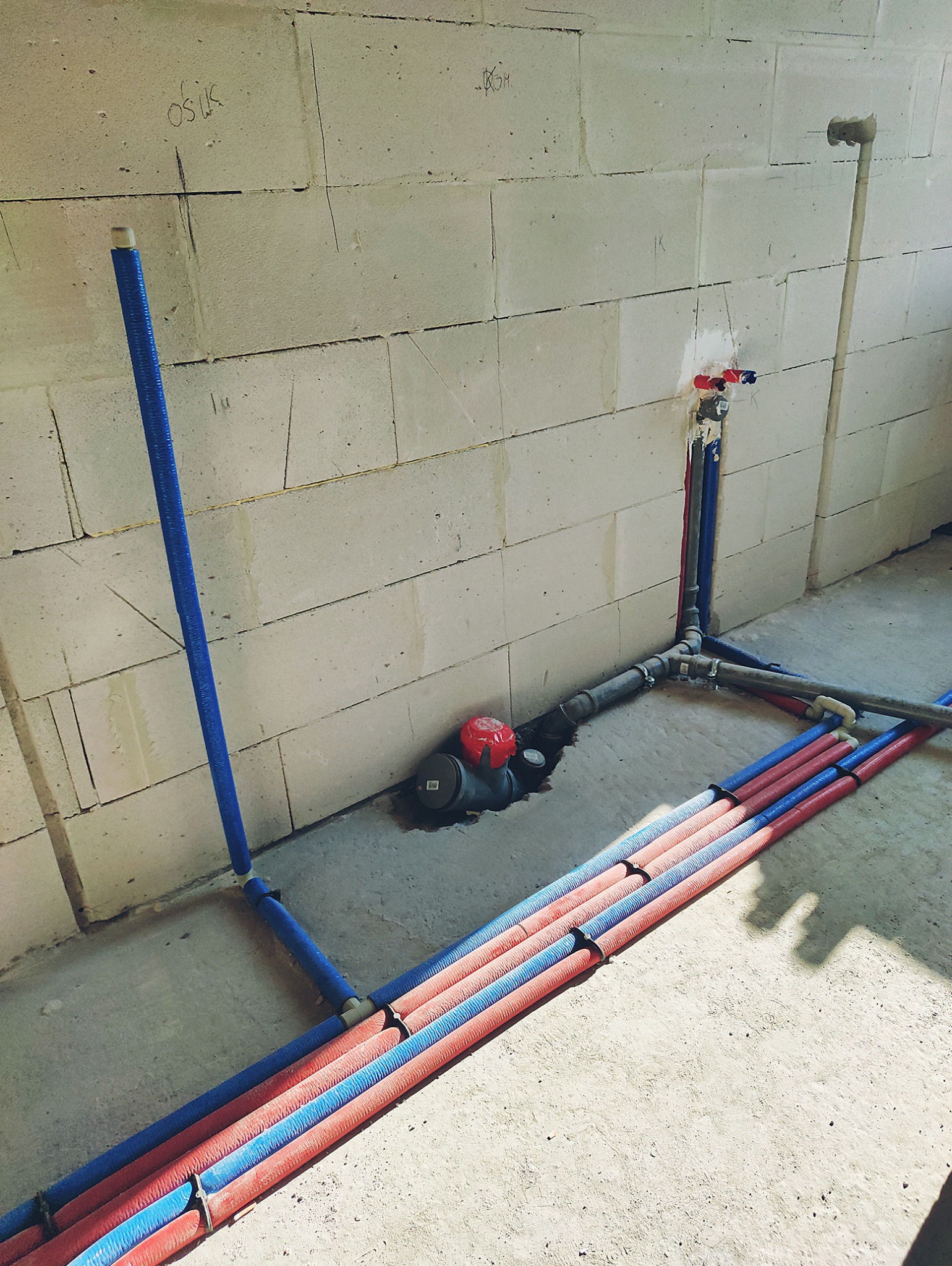 Wewnętrzna instalacja wodna i kanalizacyjna w surowym stanie, rury w izolacji karbowanej prowadzone po podłodze i ścianie z betonu komórkowego, widoczne złącza i odpowietrznik kanalizacyjny.