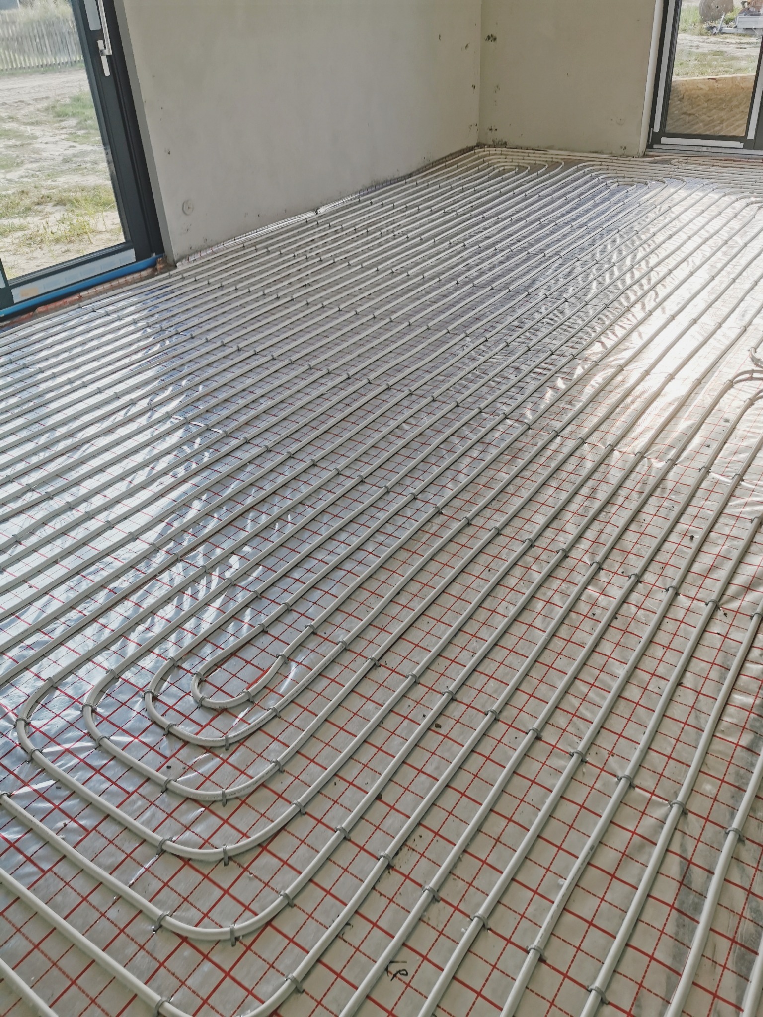 Białe rury ogrzewania podłogowego ułożone na folii z czerwoną kratką w surowym wnętrzu, widok z góry, naturalne światło wpadające przez okno.