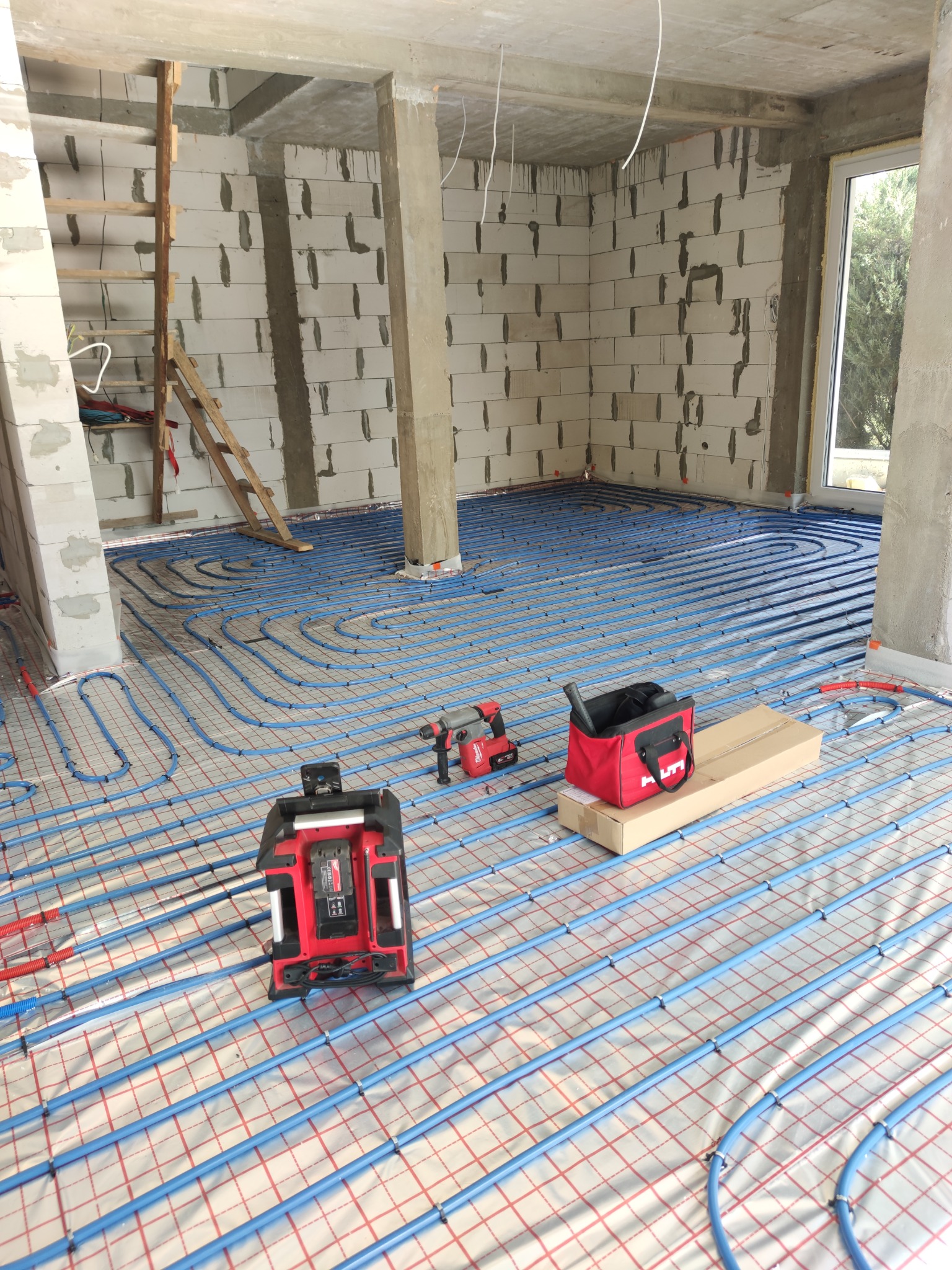 Niebieskie rury ogrzewania podłogowego ułożone na folii z czerwoną siatką w surowym wnętrzu budynku, widoczny laser krzyżowy, wiertarko-wkrętarka i torba narzędziowa.