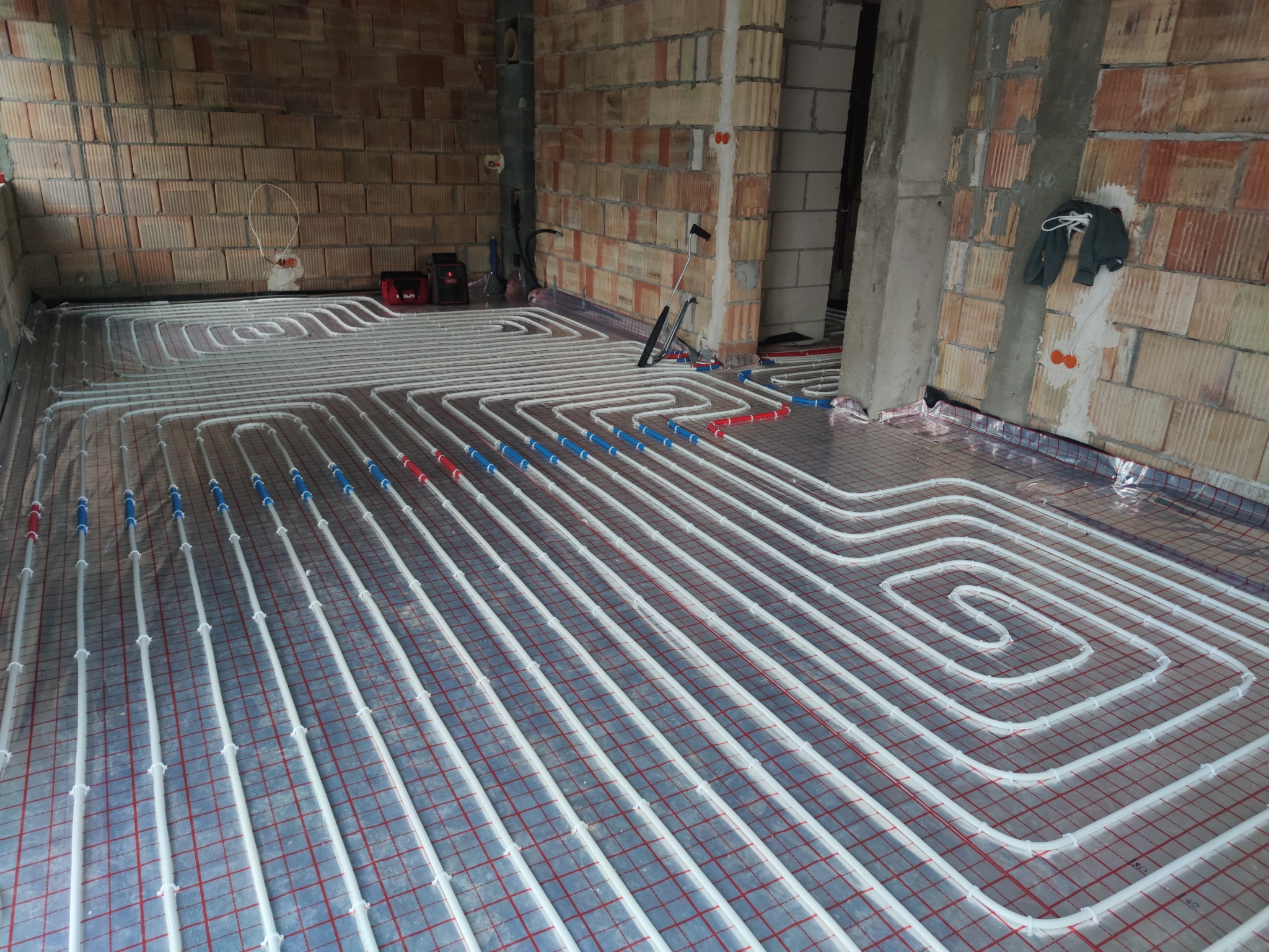 Białe rury instalacji ogrzewania podłogowego ułożone na folii z czerwoną siatką, widoczne narzędzia i surowe ściany z cegły.