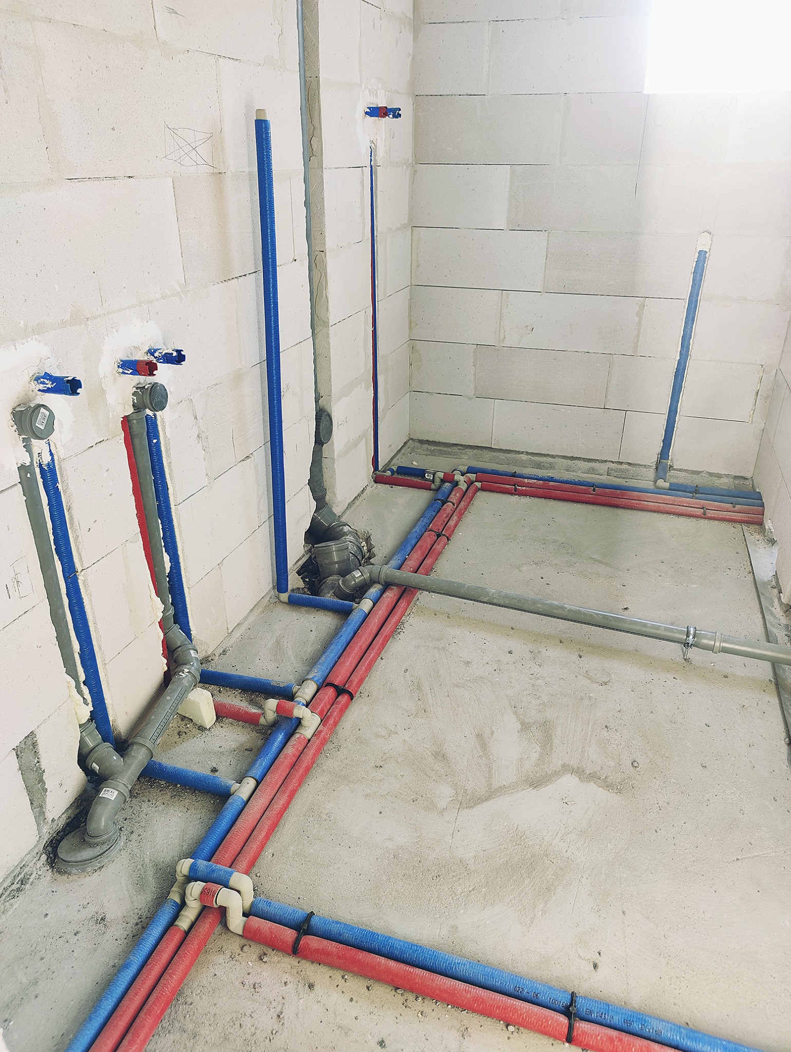 Surowy stan: Instalacja wodna i kanalizacyjna w trakcie montażu, rury czerwone i niebieskie prowadzone po podłodze i w ścianach z bloków betonowych.