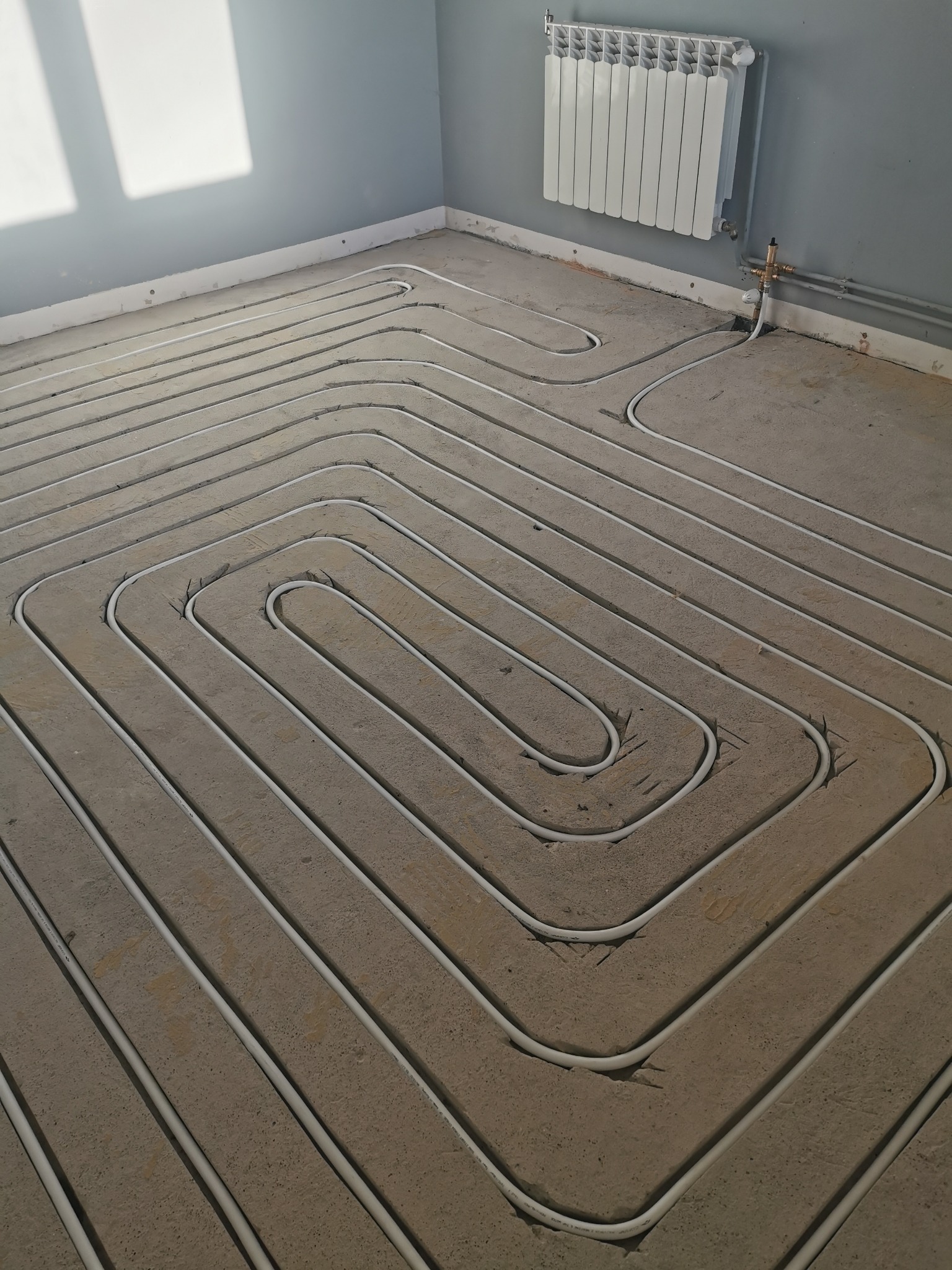 Białe rurki ogrzewania podłogowego ułożone w zygzak na szarym podłożu, z widocznym grzejnikiem na ścianie i promieniami słońca w rogu pomieszczenia.