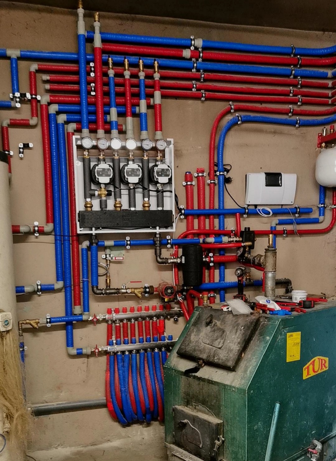 Złożona instalacja grzewcza z rurami w kolorach czerwonym i niebieskim, manometry w białej obudowie, kocioł grzewczy na pierwszym planie. Widoczne narzędzia.