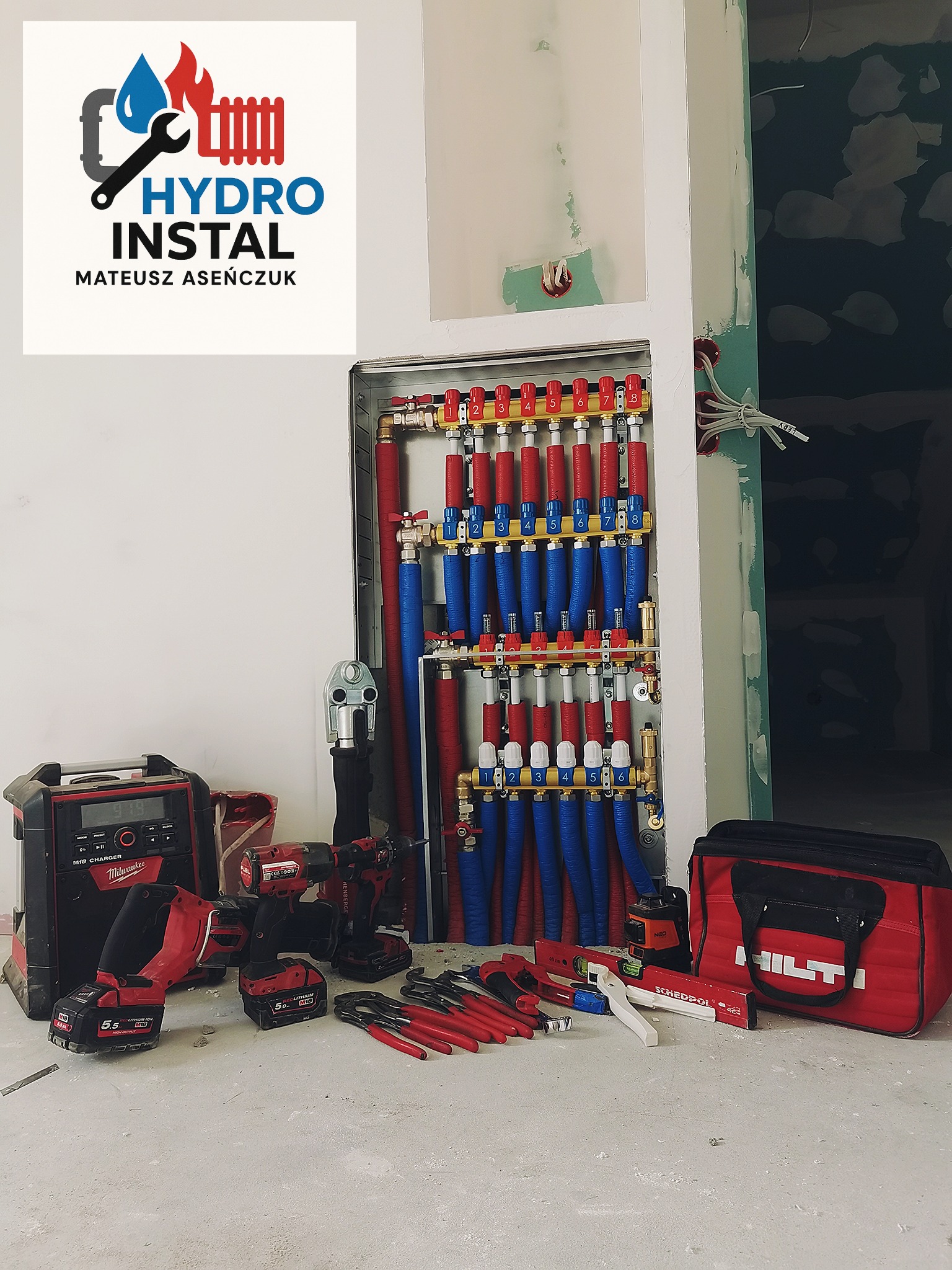 Rozdzielacz ogrzewania podłogowego w metalowej szafce, z licznymi rurami w kolorach czerwonym i niebieskim. Obok narzędzia hydrauliczne i torba HILTI. Na ścianie logo firmy.