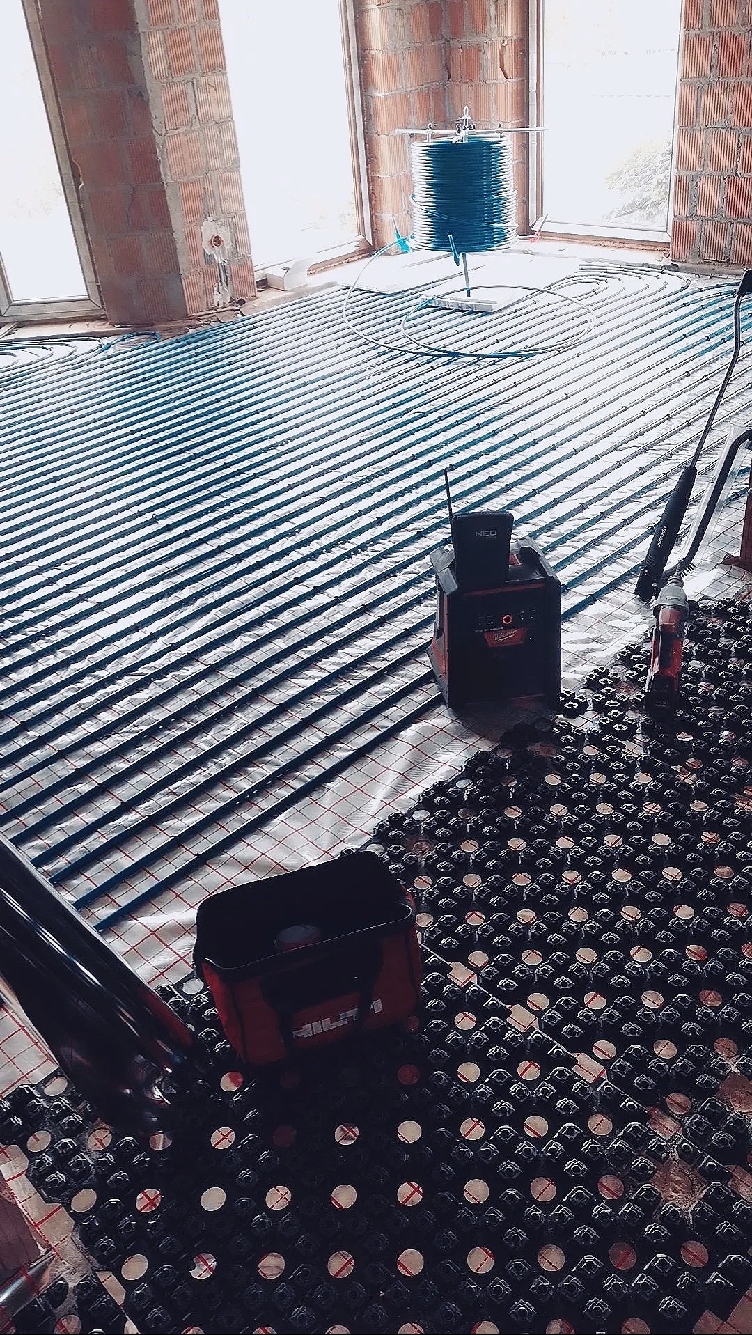 Instalacja ogrzewania podłogowego w surowym wnętrzu. Widoczne rury grzewcze, mata izolacyjna, narzędzia pracy i ściany z cegły. Etap prac przed wylewką.