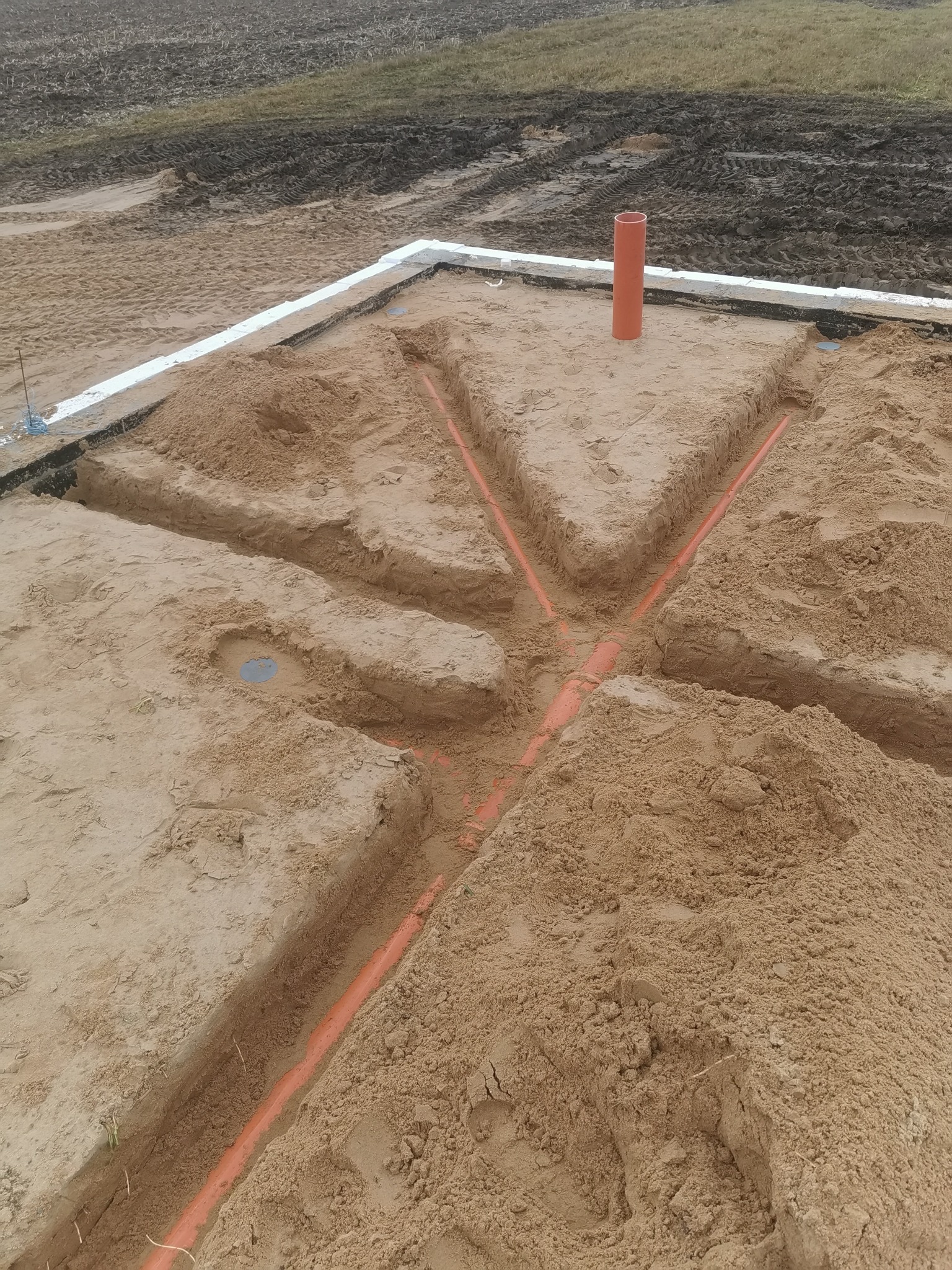 Przygotowanie podłoża pod ogrzewanie podłogowe: wykopane rowki w piasku z ułożonymi rurami grzewczymi, widoczna izolacja fundamentów i pionowa rura wentylacyjna.