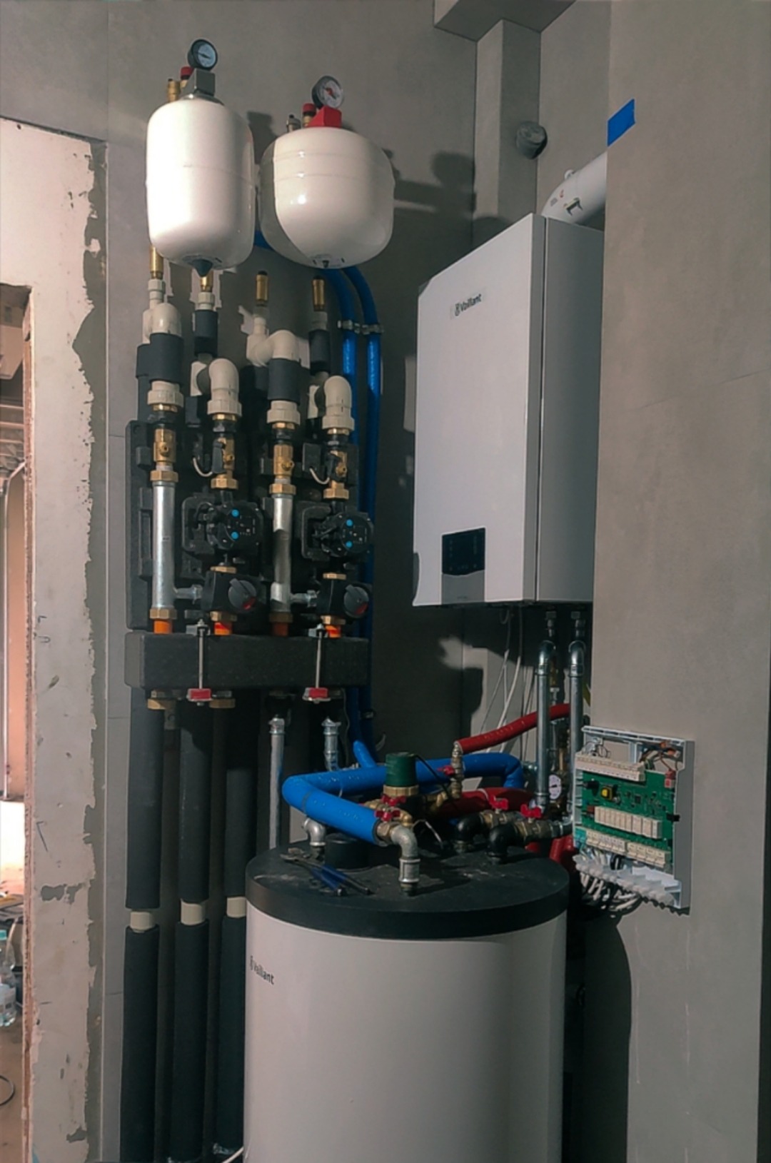 Nowoczesna kotłownia z białym piecem Vaillant, zbiornikiem i rozbudowaną instalacją hydrauliczną z czarnymi rurami w izolacji i niebieskimi rurami z tworzywa.