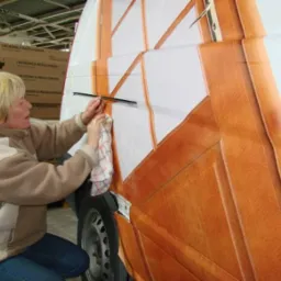 Naklejanie folii imitującej drewno na bok samochodu dostawczego przez kobietę w beżowej bluzie, widoczne pudełka w tle.