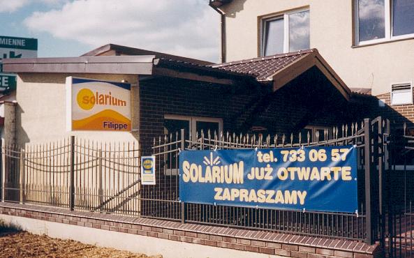 Zewnętrzny baner reklamowy solarium 'Solarium Już Otwarte' z numerem telefonu, zawieszony na metalowym ogrodzeniu przed budynkiem, obok szyldu z logo solarium 'Filippe'.