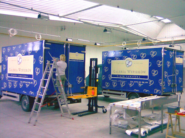 Oklejanie firmowych samochodów dostawczych reklamą 'Galeria Wypieków'. Pracownik na drabinie aplikuje folię z logo i wzorem na niebieską karoserię. W tle widoczny drugi pojazd i wózek widłowy.