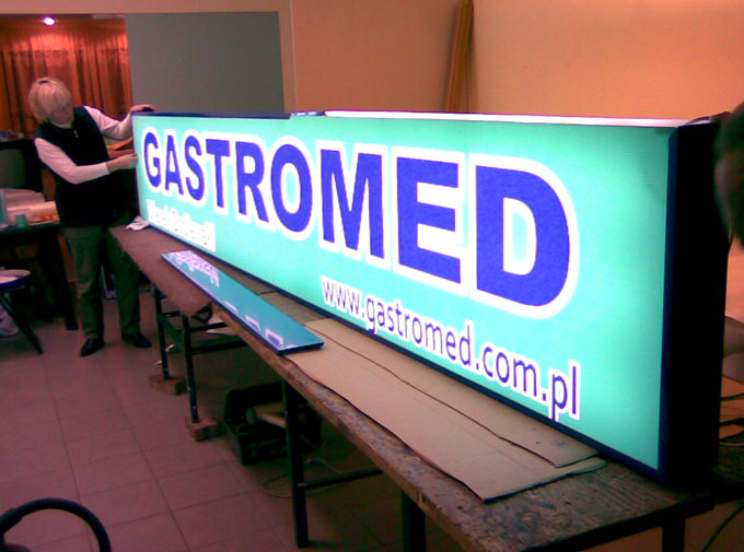Pracownik montuje podświetlany kaseton reklamowy z napisem 'GASTROMED' i adresem strony internetowej na stole roboczym.