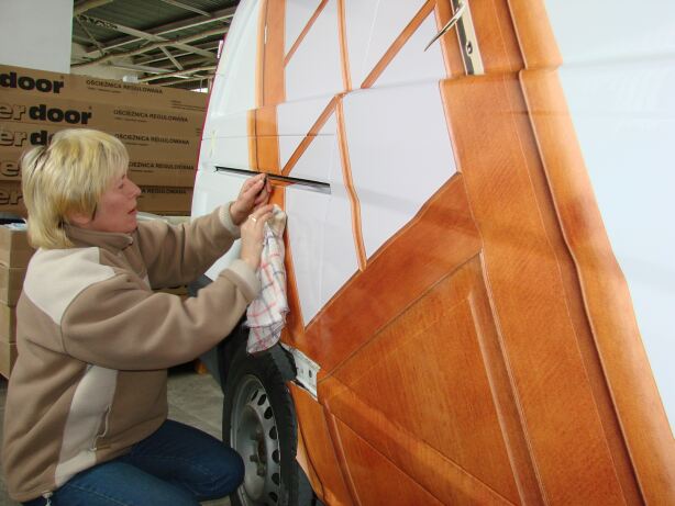 Naklejanie folii imitującej drewno na bok samochodu dostawczego przez kobietę w beżowej bluzie, widoczne pudełka w tle.