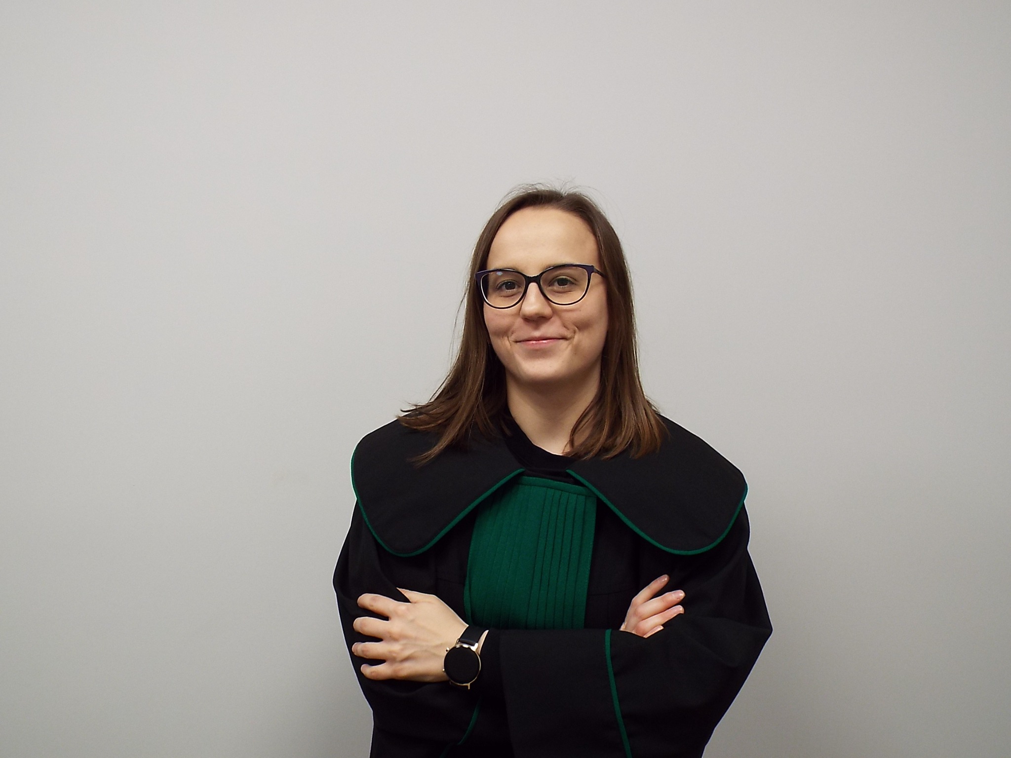 Portret uśmiechniętej kobiety w todze z zielonym żabotem i zegarkiem na ręce, skrzyżowane ręce na tle jasnej ściany.