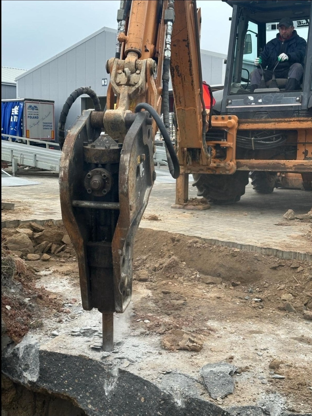 Pomarańczowa koparka z młotem hydraulicznym rozbijająca asfalt, widoczny operator w kabinie, w tle budynek przemysłowy i naczepa ciężarówki z napisem POLAND, w tle pochmurne niebo.