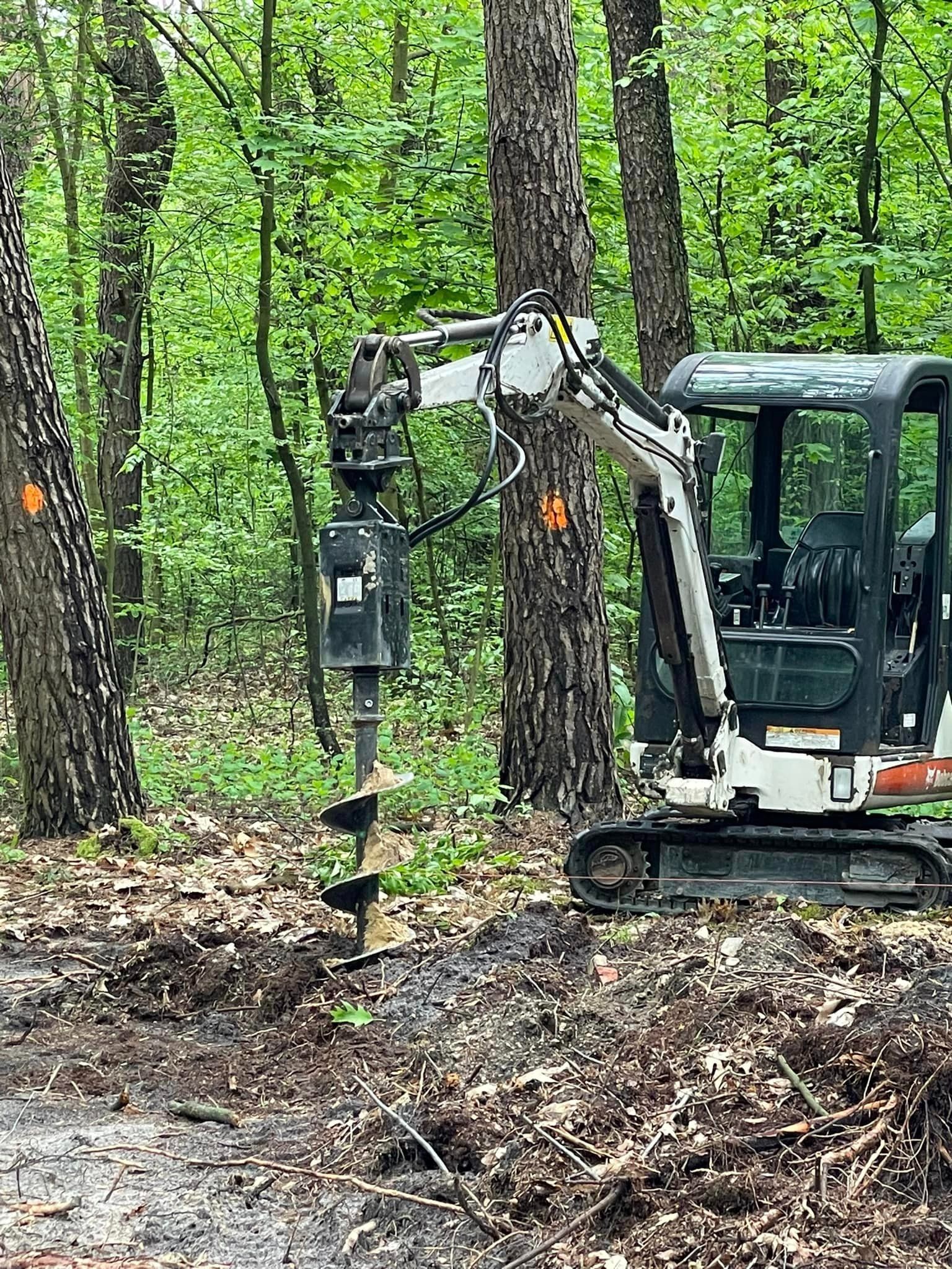 Mini koparka z wiertłem świdrowym w lesie, przygotowująca teren pod instalację, z pomarańczowymi oznaczeniami na drzewach.