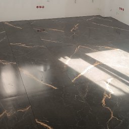 Dmytro Marynchenko - Ciemne płytki podłogowe z marmurkowym wzorem w odcieniach złota, ułożone w rogu pomieszczenia z białymi ścianami, widoczne instalacje elektryczne.