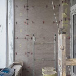 Dmytro Marynchenko - Balkon w trakcie prac glazurniczych: jasne płytki imitujące kamień, poziomica oparta o ramę okienną, drabina z wiadrami kleju i szpachelką, krzyżyki dystansowe i numeracja płytek na ścianie.