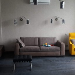 Dmytro Marynchenko - Salon w stonowanych kolorach z brązową kanapą, żółtym fotelem, czarnym pufem i ciemną podłogą; lampy ścienne z folią ochronną, zabawki na kanapie.