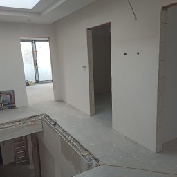 Dmytro Marynchenko - Wnętrze w trakcie szpachlowania ścian, widoczne rury instalacji grzewczej, surowe ościeżnice drzwiowe, okno z folią ochronną oraz drabina w tle.