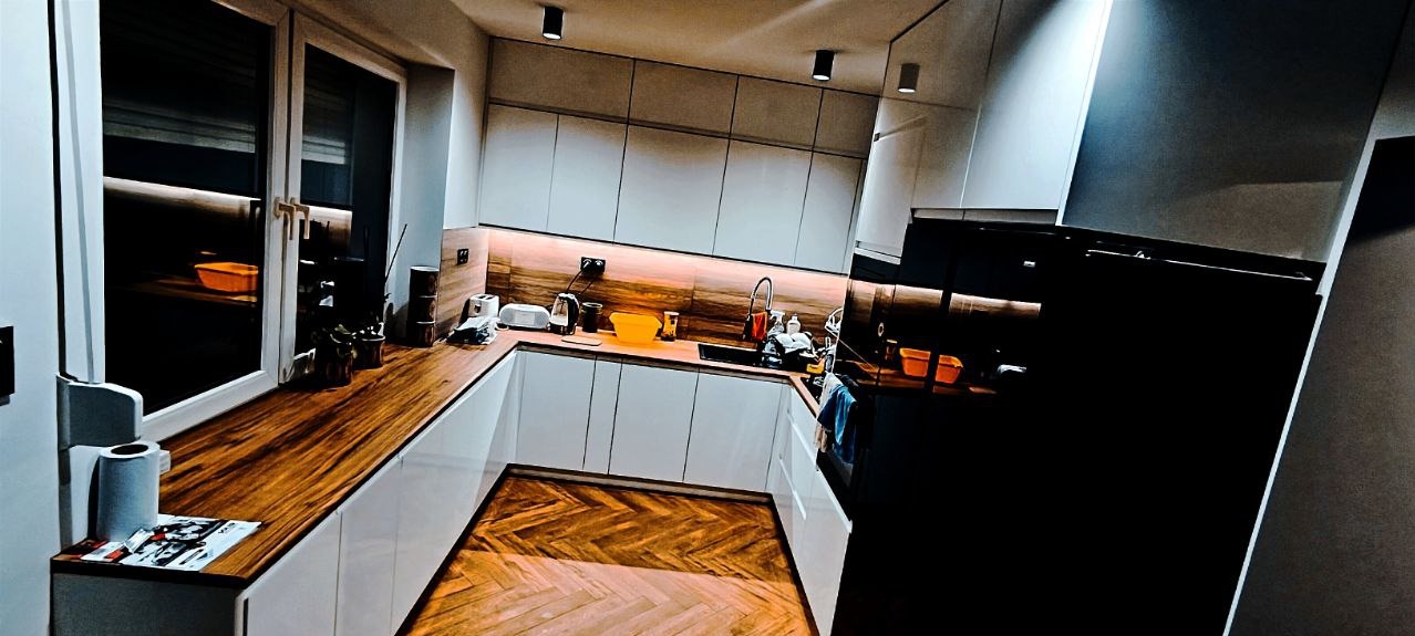 Nowoczesna kuchnia z białymi szafkami, drewnianym blatem i podłogą ułożoną w jodełkę, oświetlona punktowymi lampami sufitowymi i podświetleniem LED pod szafkami wiszącymi.