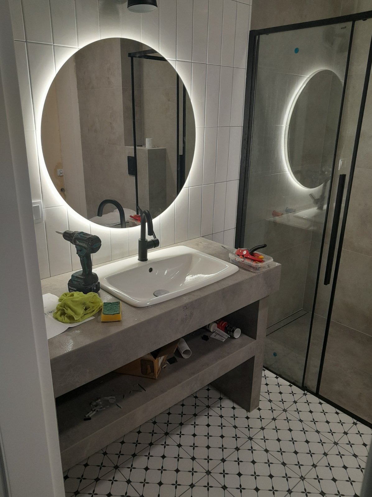 Nowoczesna łazienka w trakcie wykańczania: umywalka z czarną armaturą, podświetlane lustro, kabina prysznicowa z czarnymi profilami, widoczne narzędzia i akcesoria glazurnicze na blacie umywalkowym.