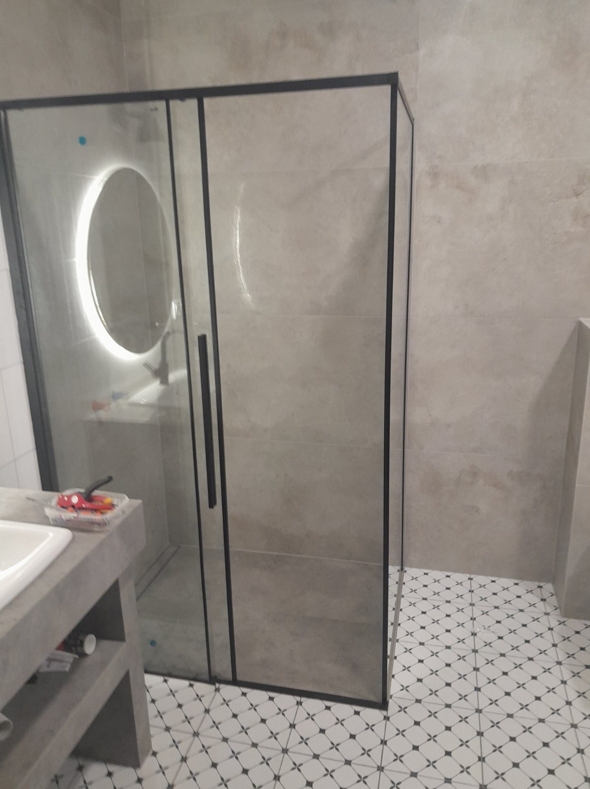 Nowoczesna kabina prysznicowa z czarnymi profilami i okrągłym lustrem z podświetleniem LED, widoczna umywalka z betonowym blatem i wzorzysta podłoga.