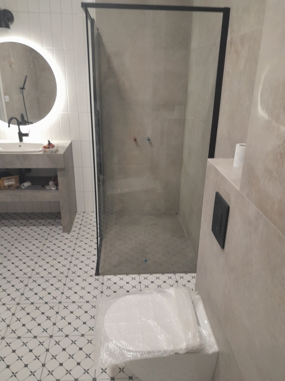 Nowoczesna łazienka w trakcie wykańczania: kabina prysznicowa z czarną ramą, umywalka z okrągłym lustrem podświetlanym LED, biało-czarna mozaika podłogowa, toaleta zabezpieczona folią.