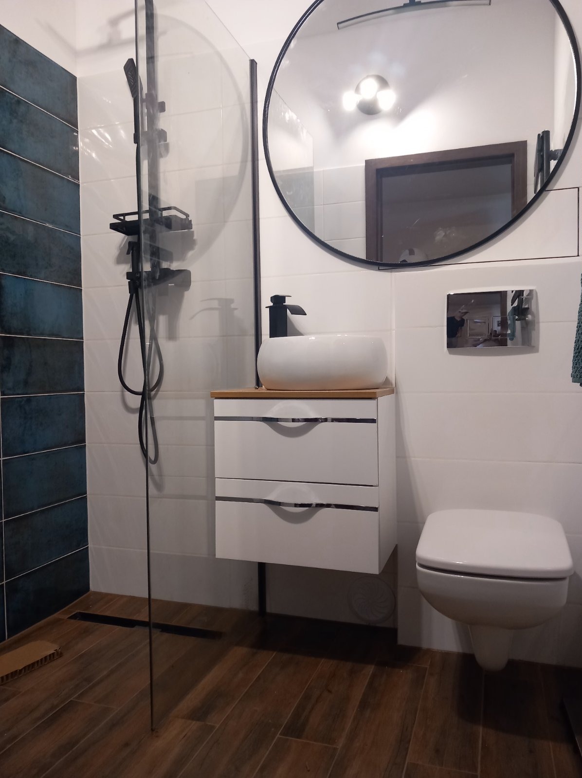 Nowoczesna łazienka z kabiną prysznicową typu walk-in, umywalką nablatową na szafce z dwiema szufladami, okrągłym lustrem w czarnej ramie i podwieszaną toaletą.