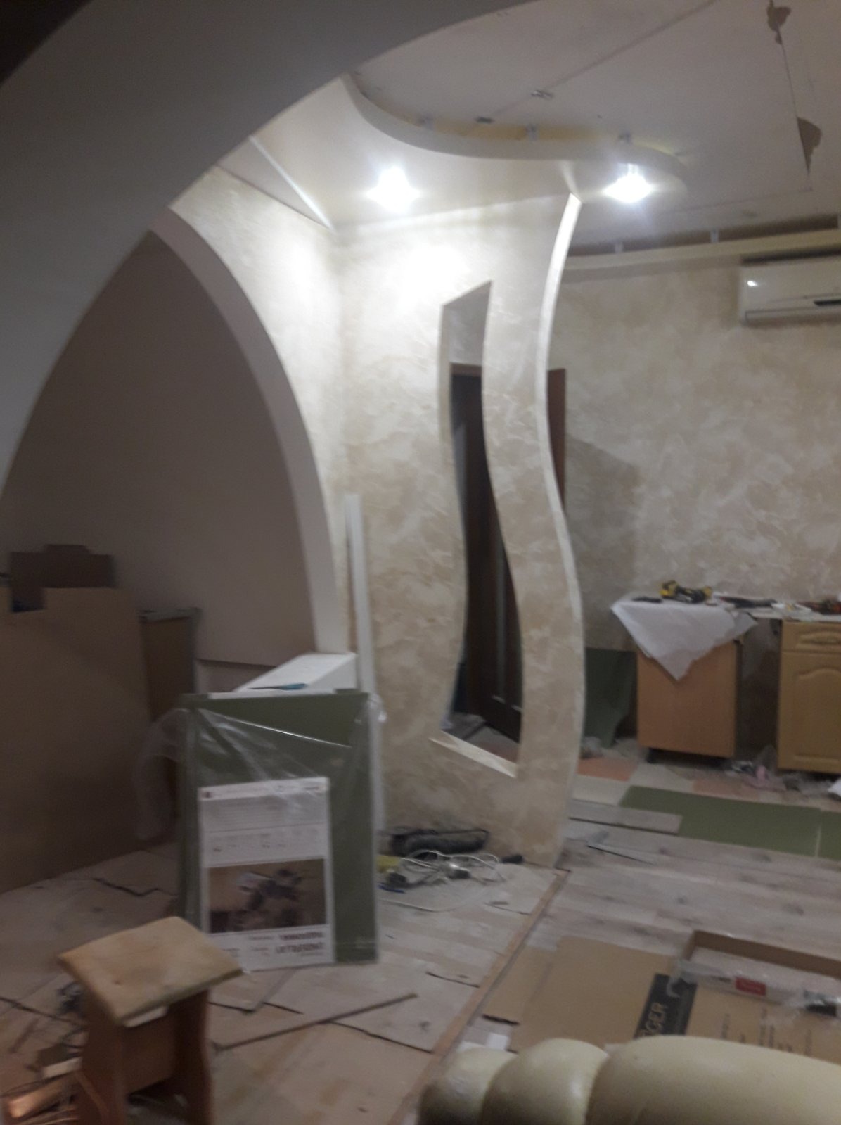 Niedokończone wnętrze ze ścianami pokrytymi dekoracyjnym tynkiem, widoczne elementy konstrukcyjne łuków i niestandardowego otworu drzwiowego, podłoga częściowo zabezpieczona kartonem, w tle meble...