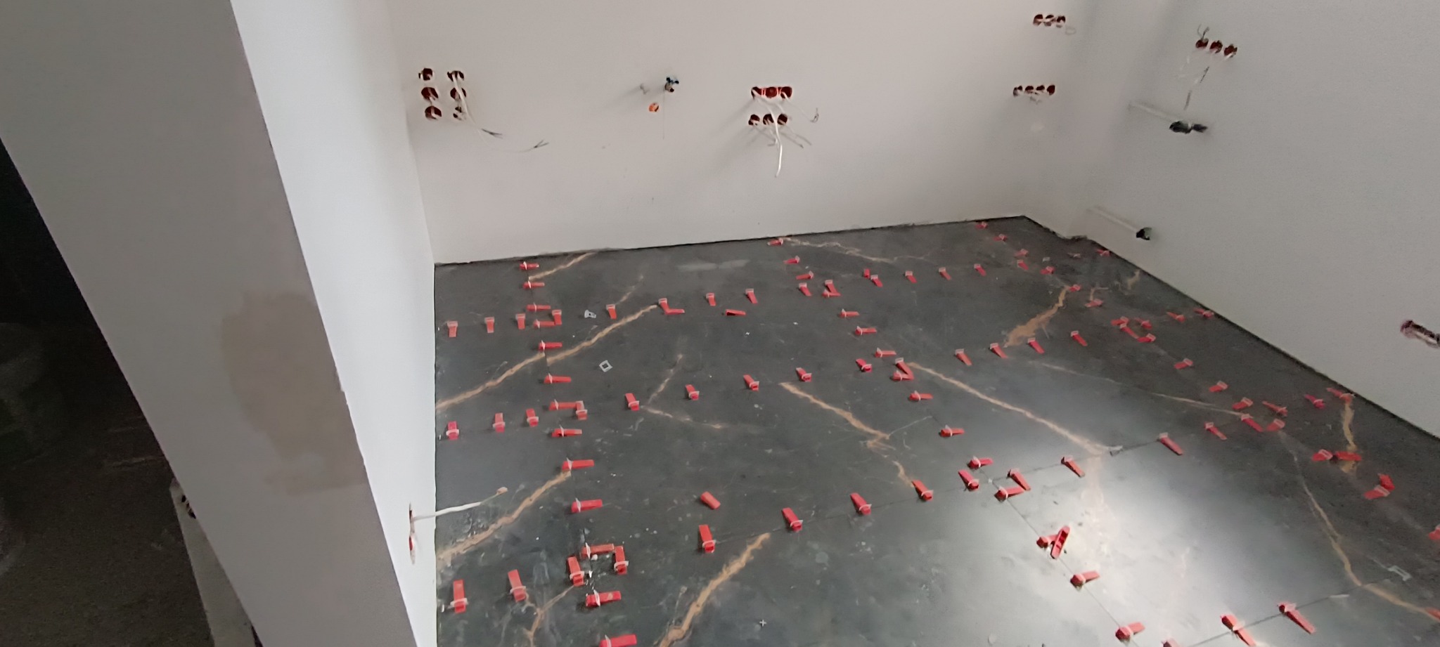 Podłoga w trakcie układania płytek imitujących marmur z użyciem czerwonych klipsów poziomujących, widoczne przygotowane punkty elektryczne na ścianach.