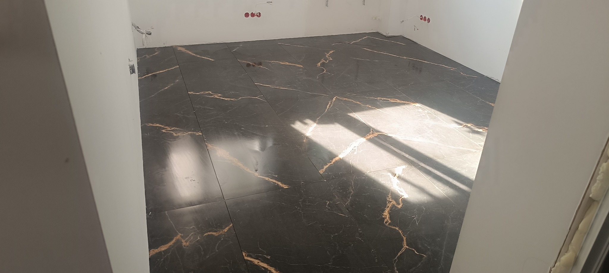 Ciemne płytki podłogowe z marmurkowym wzorem w odcieniach złota, ułożone w rogu pomieszczenia z białymi ścianami, widoczne instalacje elektryczne.