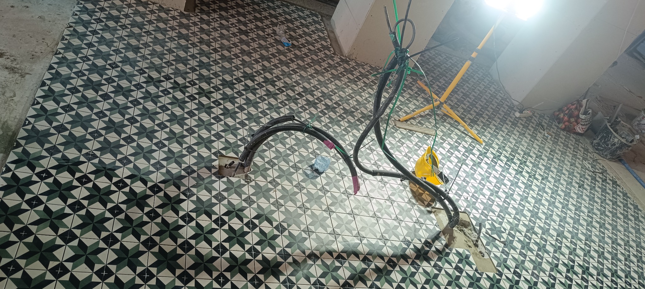 Układanie płytek podłogowych w łazience z wzorem geometrycznym gwiazd, widoczne kable elektryczne i lampa robocza.