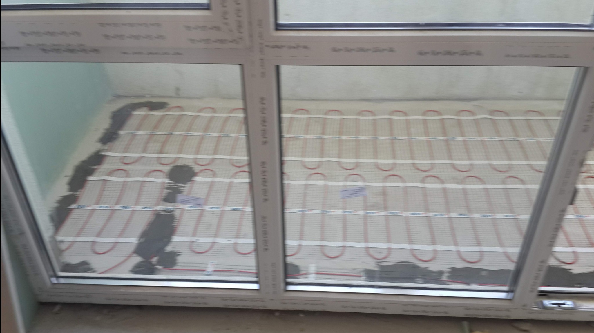 Instalacja ogrzewania podłogowego elektrycznego w trakcie zalewania wylewką, widoczne kable grzewcze i fragment okna.