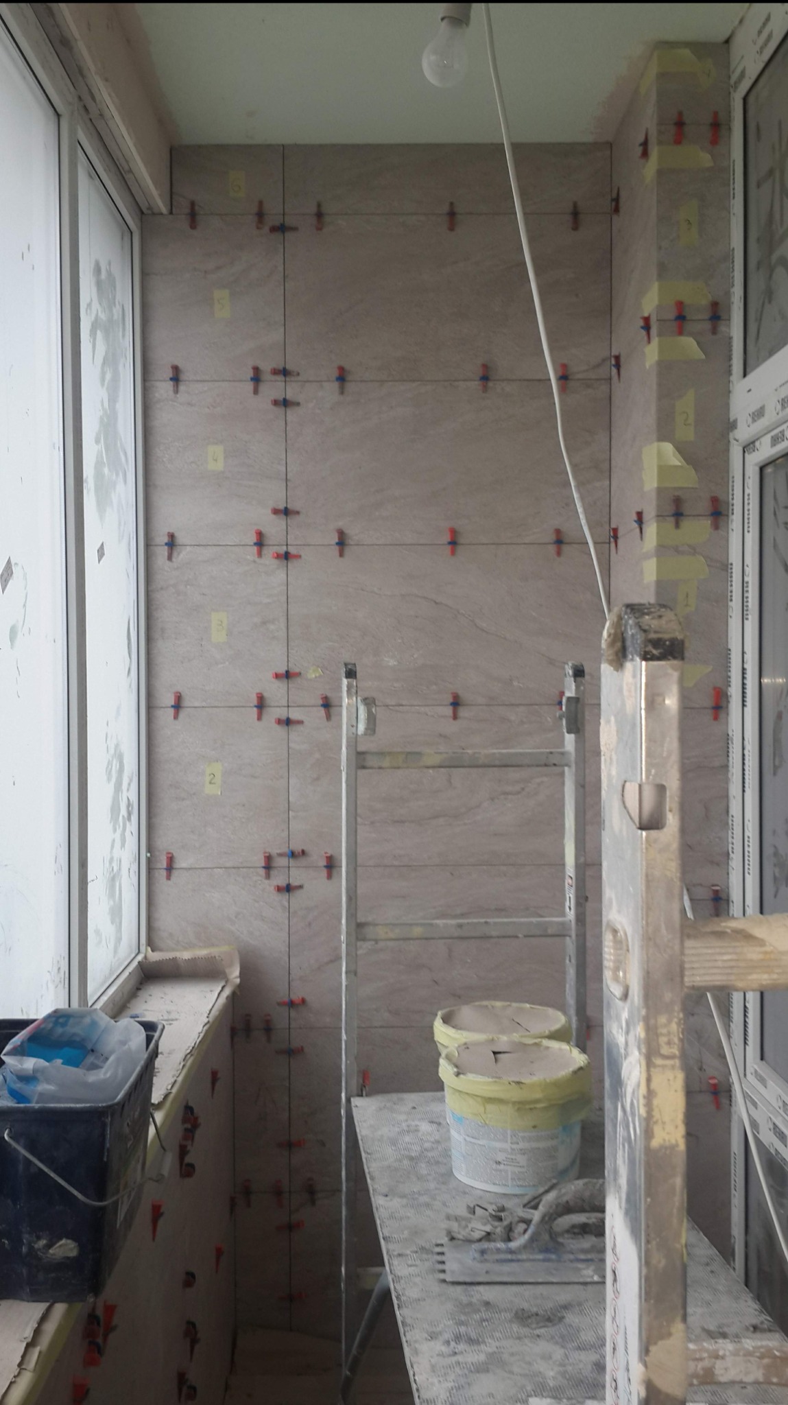 Balkon w trakcie prac glazurniczych: jasne płytki imitujące kamień, poziomica oparta o ramę okienną, drabina z wiadrami kleju i szpachelką, krzyżyki dystansowe i numeracja płytek na ścianie.