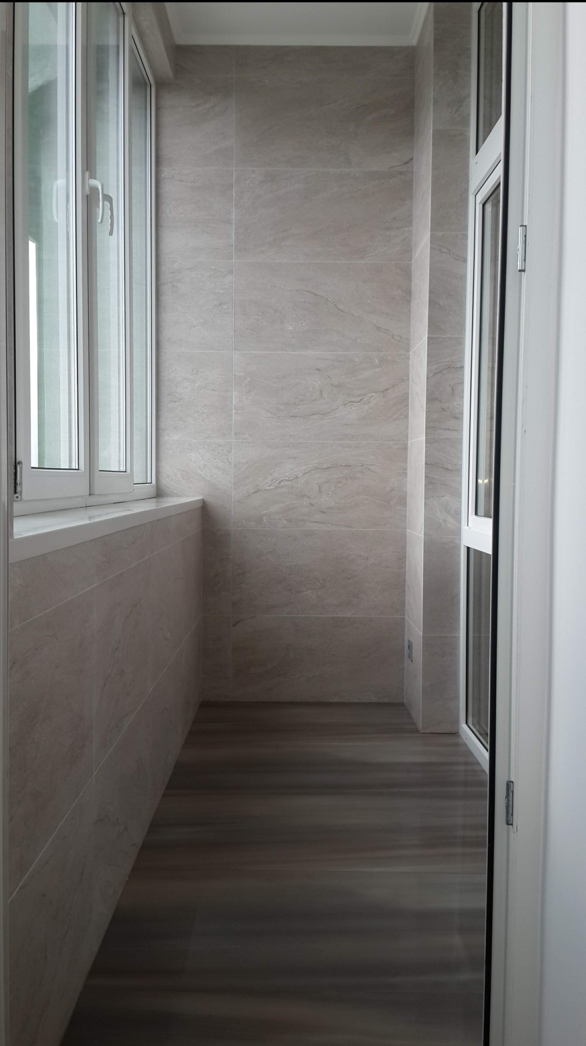 Wnętrze wykończonego balkonu z szarymi płytkami imitującymi marmur na ścianach i podłodze, białe okna i drzwi balkonowe. Widok z perspektywy wnętrza mieszkania.