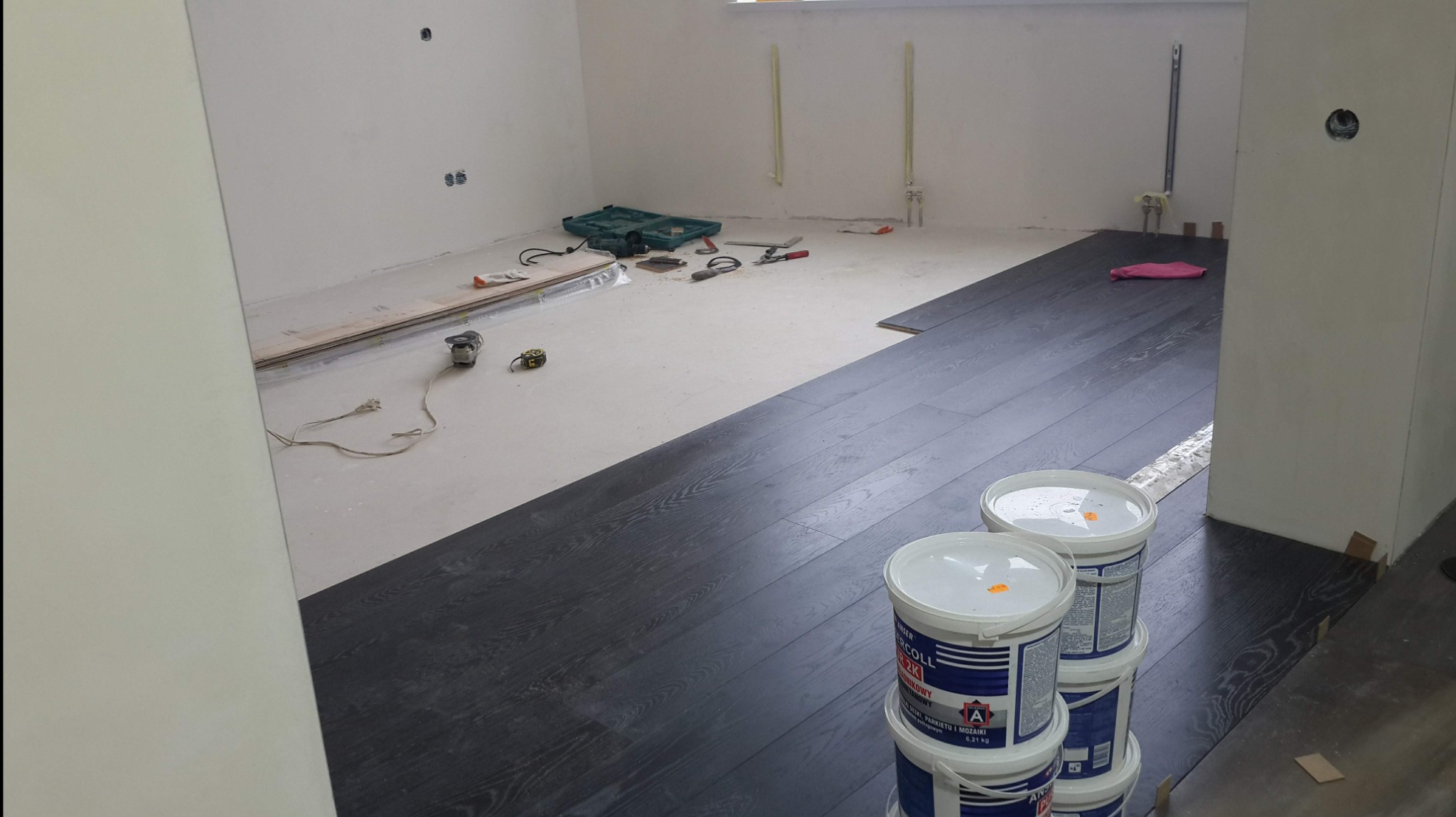 Częściowo ułożony parkiet z ciemnego drewna w pomieszczeniu w trakcie remontu, widoczne narzędzia, klej do parkietu w wiadrach i przygotowane deski.