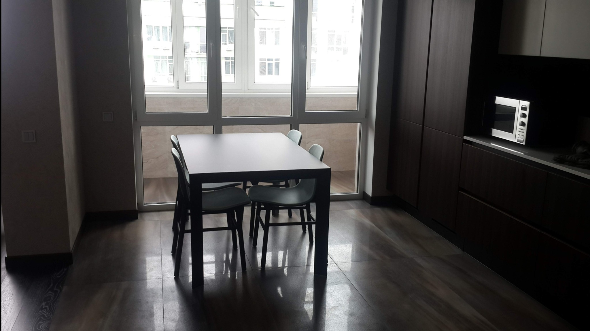 Nowoczesna jadalnia z ciemnymi meblami, płytkami podłogowymi imitującymi drewno i dużym oknem balkonowym w jasnych ramach.