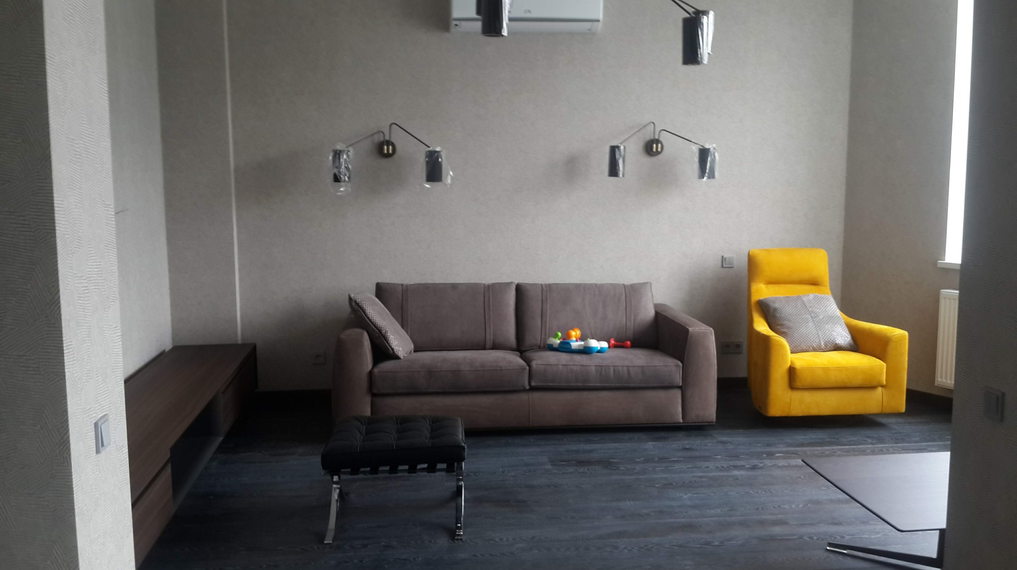 Salon w stonowanych kolorach z brązową kanapą, żółtym fotelem, czarnym pufem i ciemną podłogą; lampy ścienne z folią ochronną, zabawki na kanapie.