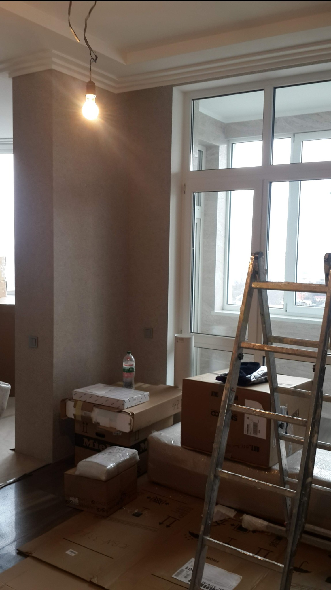Pokój w trakcie remontu: widoczne kartony z materiałami budowlanymi, drabina oparta o okno, surowa żarówka zwisająca z sufitu z dekoracyjną sztukaterią.
