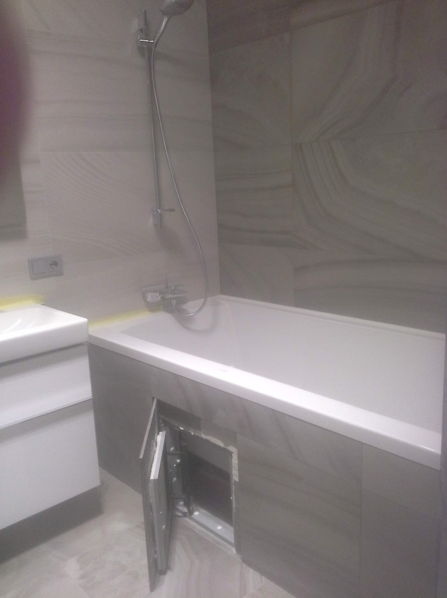 Nowoczesna łazienka z wanną obudowaną płytkami imitującymi kamień, białą umywalką i otwartym panelem rewizyjnym pod wanną.