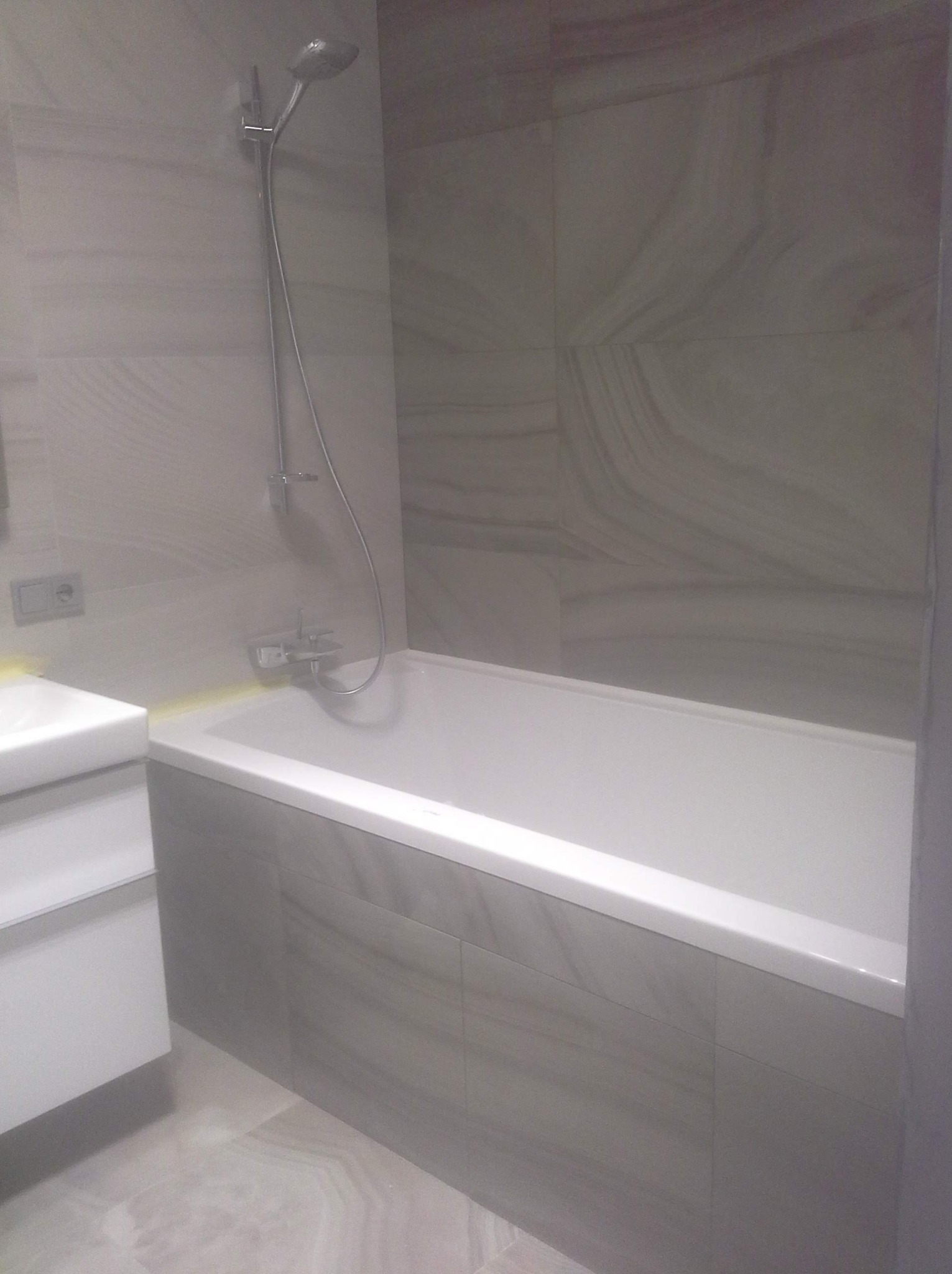 Biała wanna z panelem z płytek imitujących kamień, bateria natryskowa i umywalka w nowoczesnej łazience we Wrocławiu, wykończonej w odcieniach szarości.