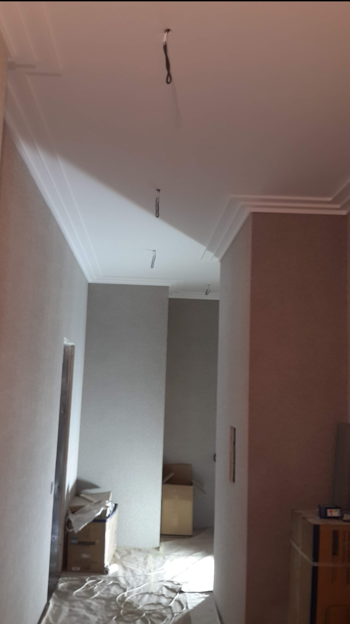 Korytarz w trakcie remontu, widoczne szpachlowane ściany, listwy sufitowe i kable zwisające z sufitu, podłoga zabezpieczona folią malarską.