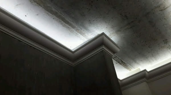 Narożnik sufitu z dekoracyjną listwą oświetleniową LED, surowy tynk na ścianach i suficie, widoczne ślady po szpachlowaniu, stan surowy.