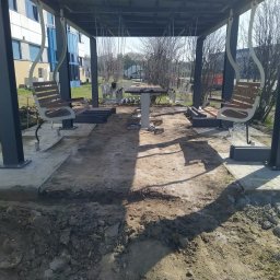 Bruk-tor - Montaż altany z huśtawkami: betonowe fundamenty, stalowe słupy, drewniane siedziska. Widoczne prace ziemne i przygotowanie terenu pod konstrukcję.