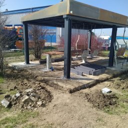 Bruk-tor - Budowa altany z huśtawkami i stołem na betonowej podstawie. Przygotowane obrzeża z kostki, teren częściowo rozkopany. W tle budynek przemysłowy i pojazd.