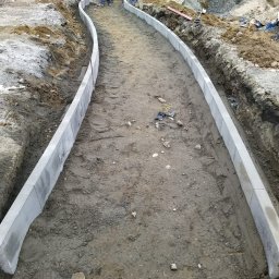 Bruk-tor - Zakrzywiony chodnik z szarej kostki obrzeżowej w trakcie budowy, widoczne ślady opon i ziemia. W tle zaparkowane samochody i osoba w odblaskowej kamizelce.