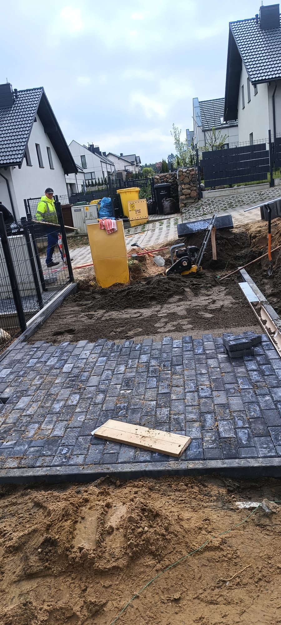 Fragment układanej nawierzchni z szarej kostki brukowej na podsypce piaskowej, widoczny pracownik w odblaskowej kamizelce oraz narzędzia i maszyny budowlane w tle.